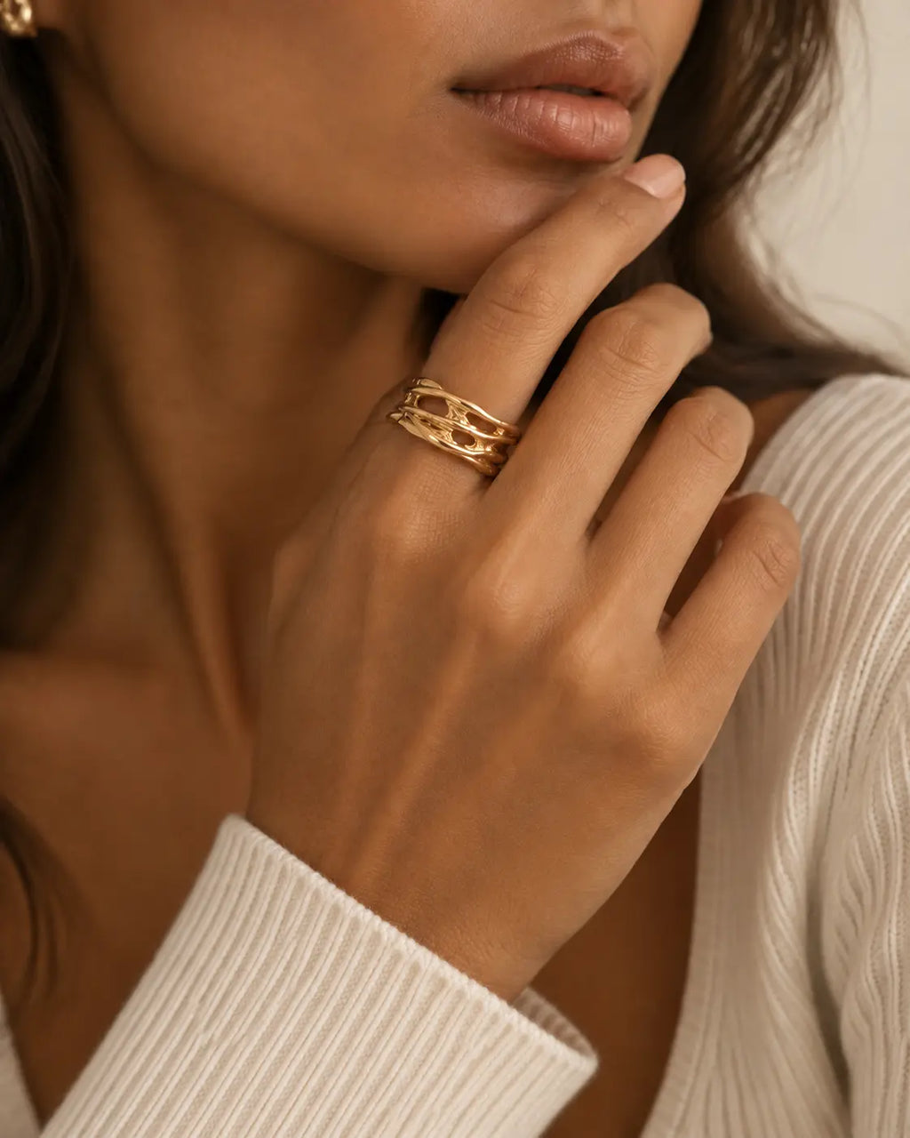 Gold-tone open ring with raised rails and irregular cutouts, worn on a hand near the chin.