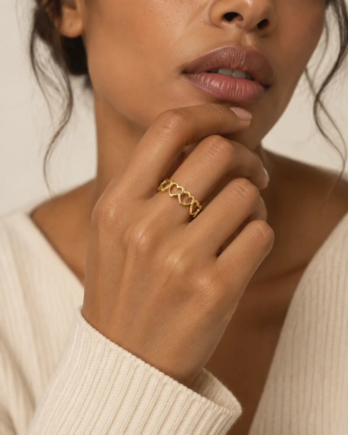 Gold-tone open band ring with linked heart outlines, worn on a hand near the chin.