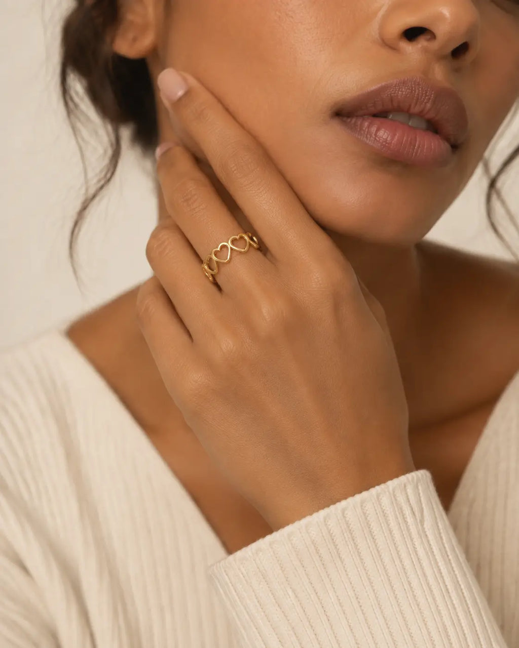 Gold-tone open band ring with linked heart outlines, worn with a finger resting beside the cheek.