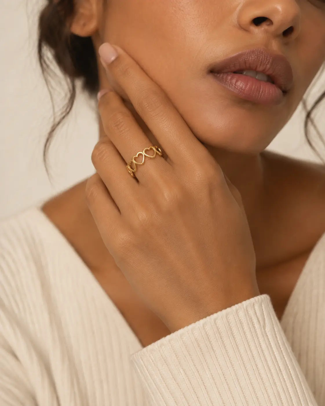 Gold-tone open band ring with linked heart outlines, worn with a finger resting beside the cheek.