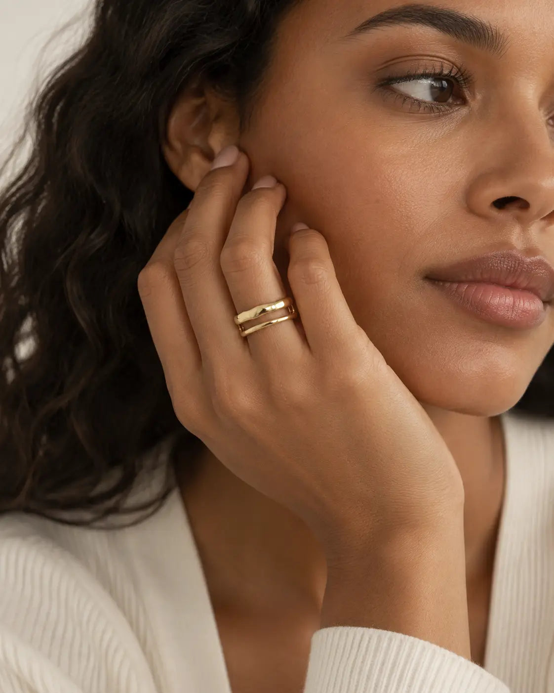 Gold-tone ring with two uneven connected bands, worn with a hand beside the cheek.