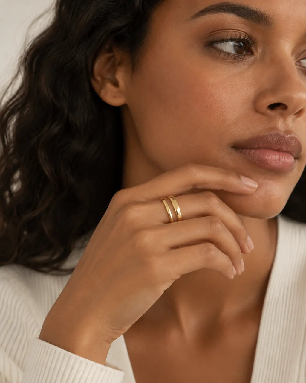 Gold-tone ring with two uneven connected bands, worn on a hand below the chin.