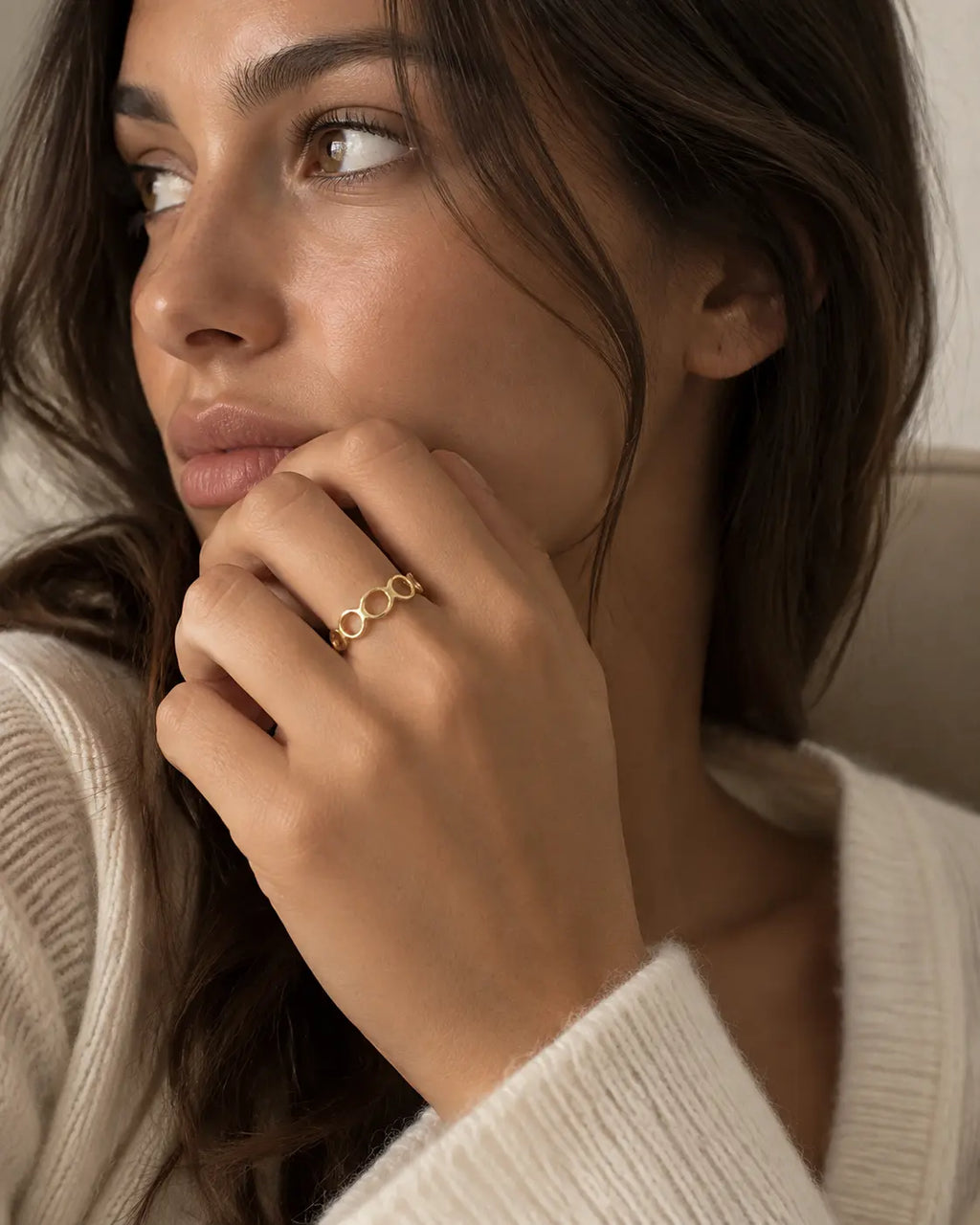 Gold-tone open band ring with linked geometric loops, worn on a hand near the chin.