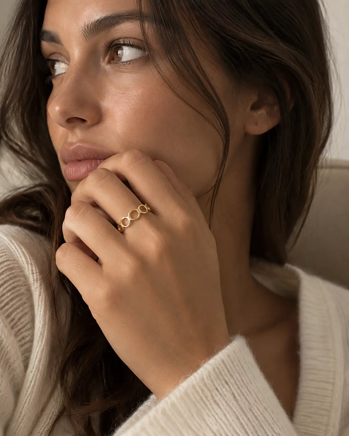 Gold-tone open band ring with linked geometric loops, worn on a hand near the chin.
