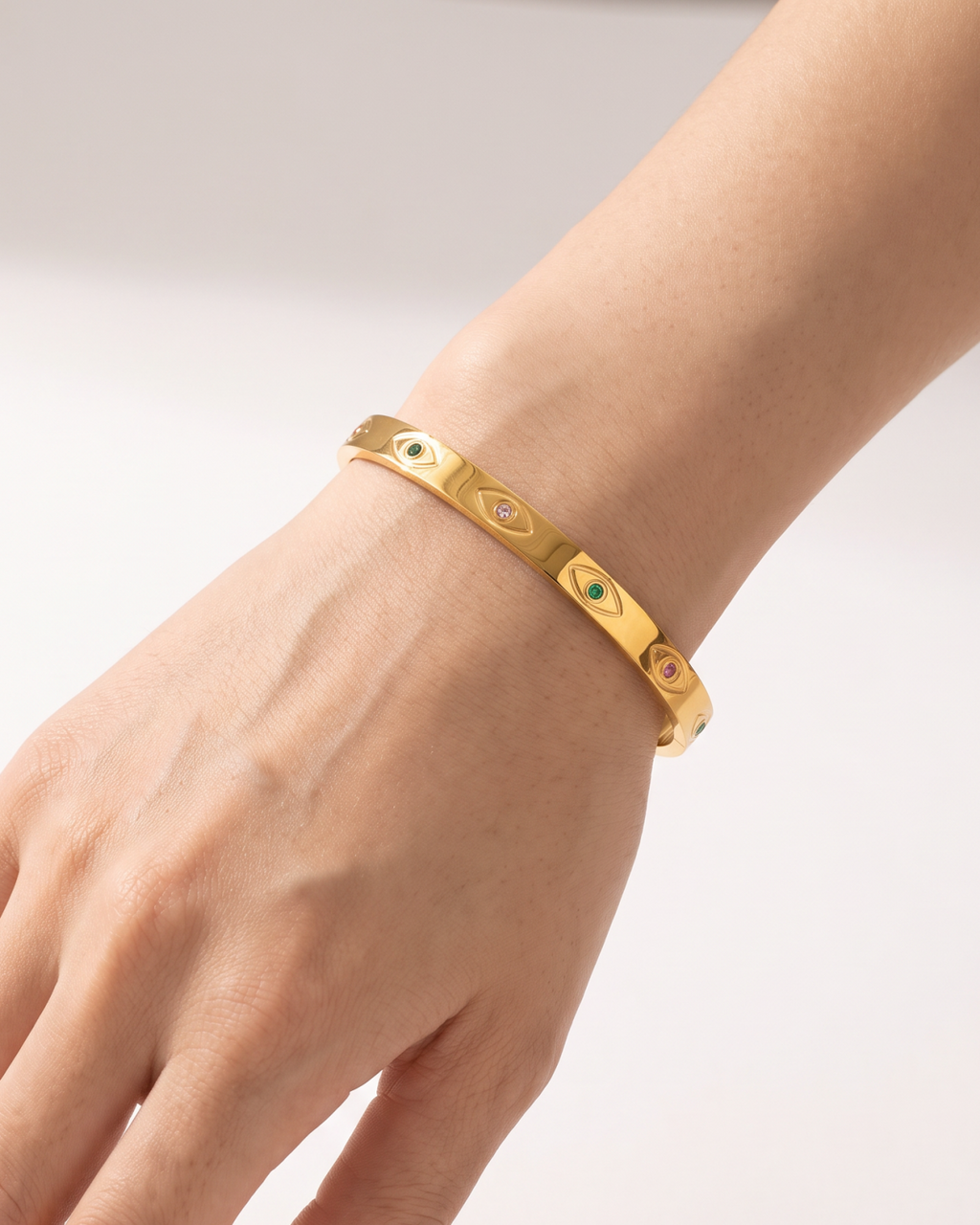 Gold-tone bangle with eye motifs and coloured centres, worn with the hand angled down.