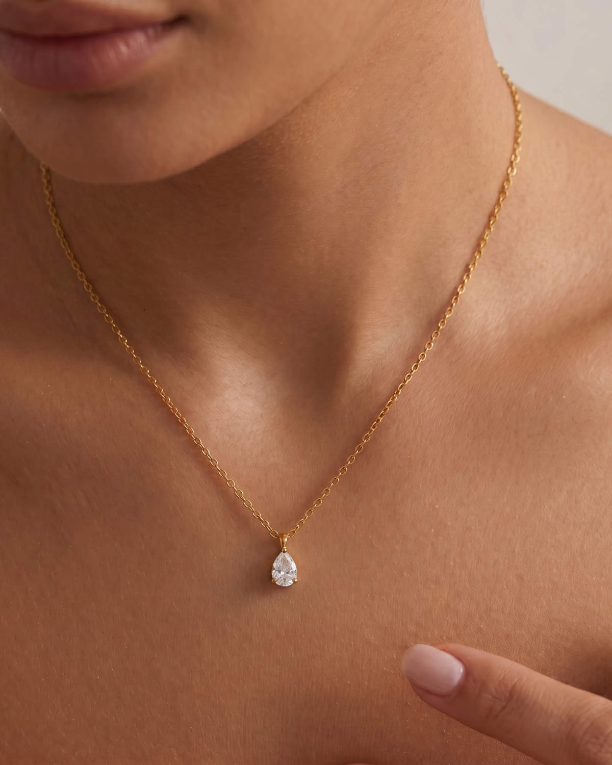 Gold-tone teardrop pendant with a clear stone accent on a fine chain, worn at a V-shaped neckline.
