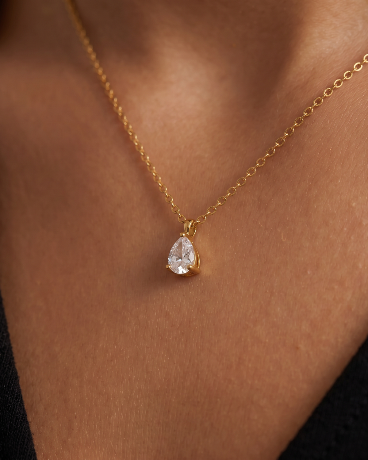 Gold-tone teardrop pendant with a clear stone accent on a fine chain, close-up at the collarbone.